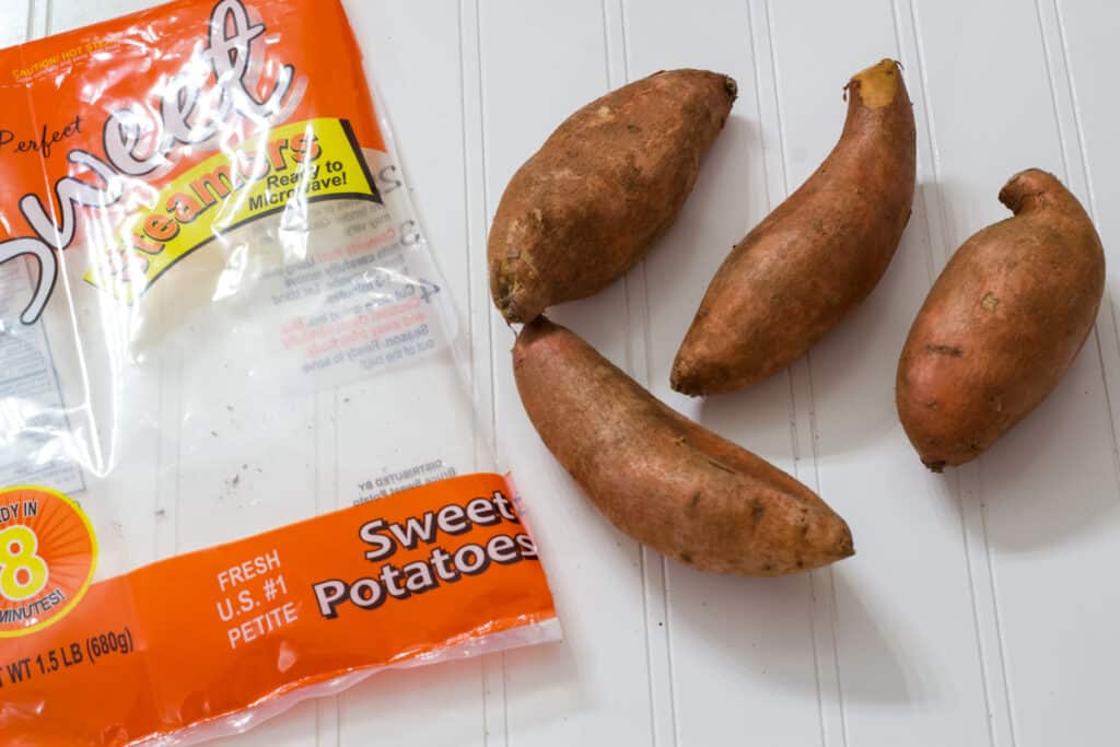 Four whole uncooked sweet potatoes next to the empty bag that shows that they are petite sweet potatoes.