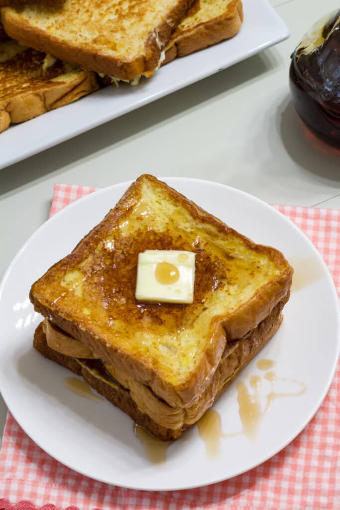 Three slices of french toast on a white plate and a plate with many pieces of french toast on it in the background.
