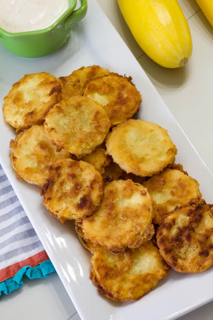 A white plate of cooked breaded summer squash and a bowl of ranch dressing next to it.