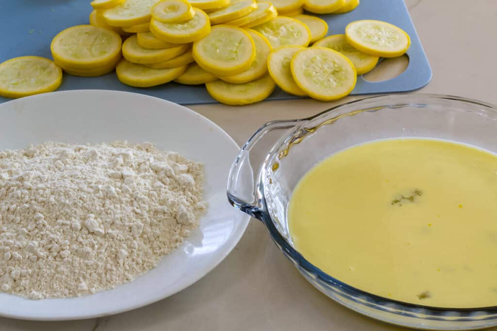 A plate of flour and glass pie pan with the egg mixture in the foreground and some sliced squash in the background.