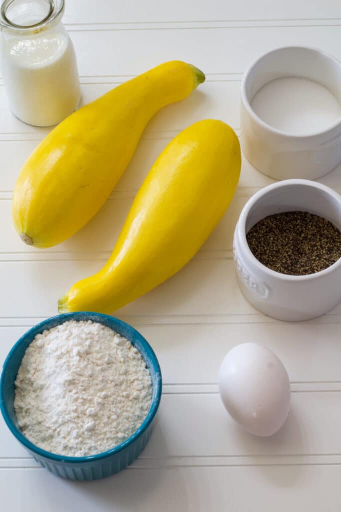 Two yellow squash, flour, milk, an egg, salt and pepper laying on a table.