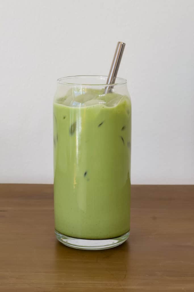 One tall glass of iced matcha tea latte sitting on a wooden countertop.