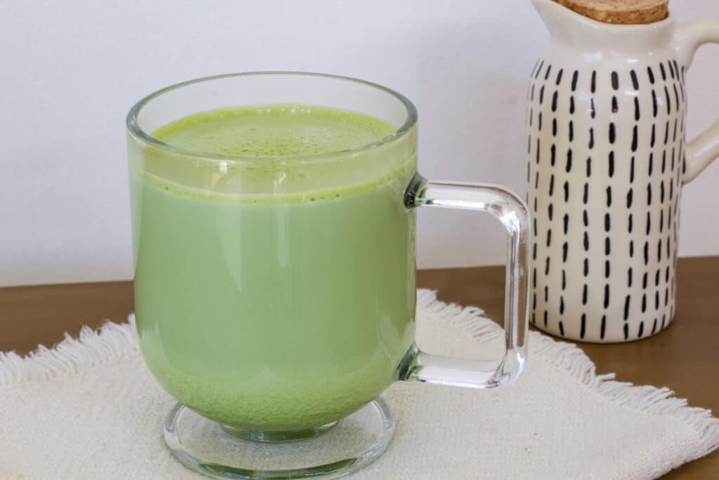 One glass coffee mug full of hot matcha tea latte, there is a small ceramic jug next to it.