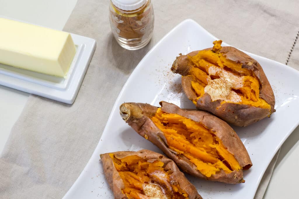 Three cooked sweet potatoes on a white plate with a stick of butter and shaker full of cinnamon next to it.