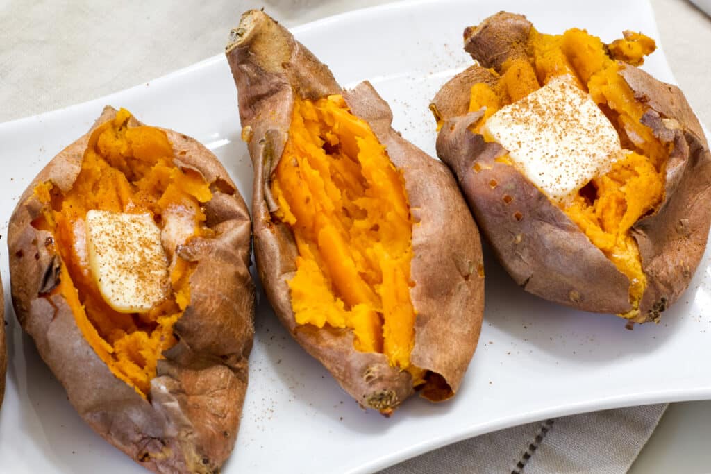 Three baked sweet potatoes on a white plate, two have butter and cinnamon and one is plain.