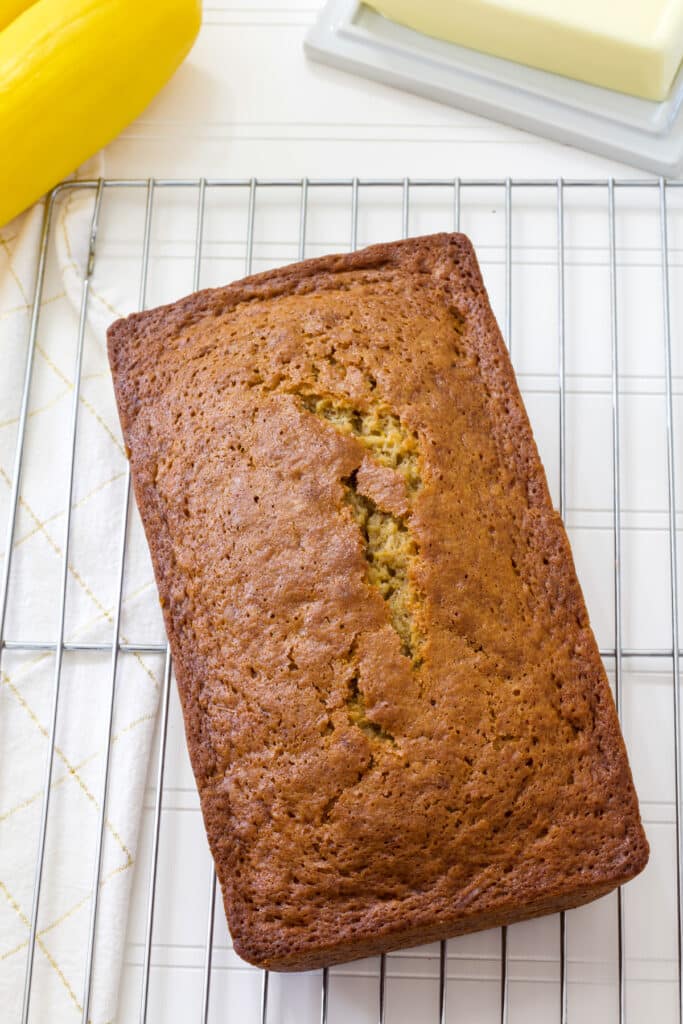 The entire uncut loaf sitting on a wire rack, there is a stick of butter and whole yellow squash partially visible nearby.