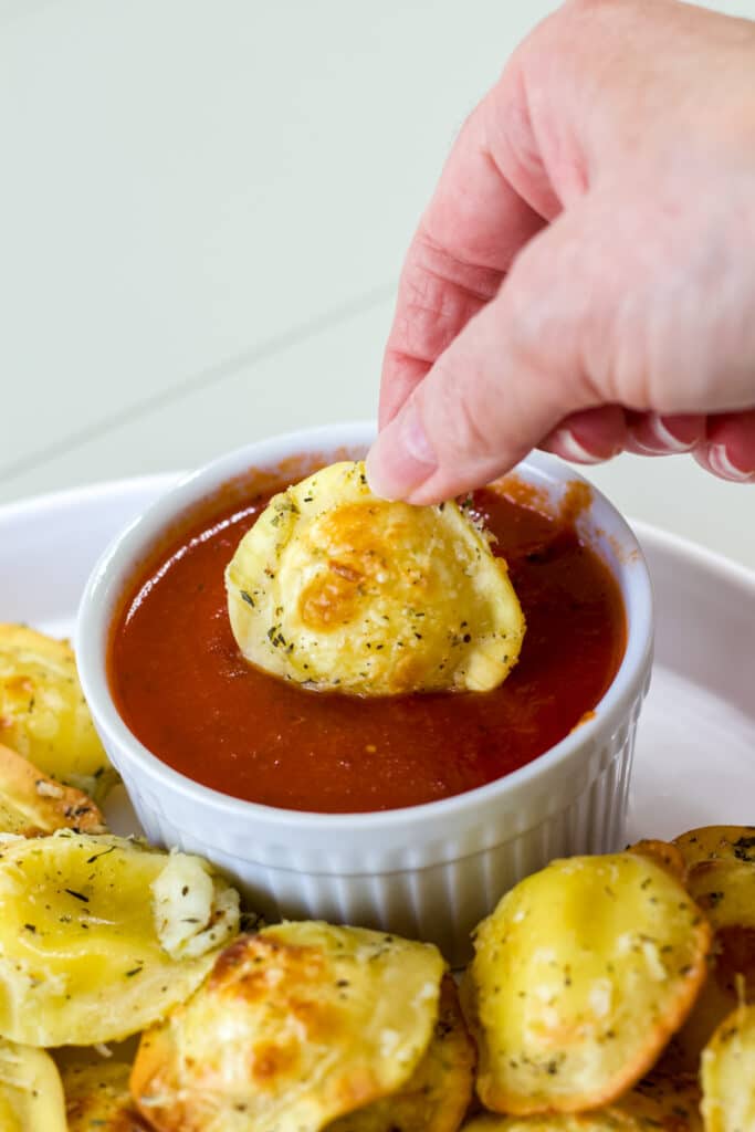 A hand dipping one fried ravioli into a small bowl of dipping sauce.