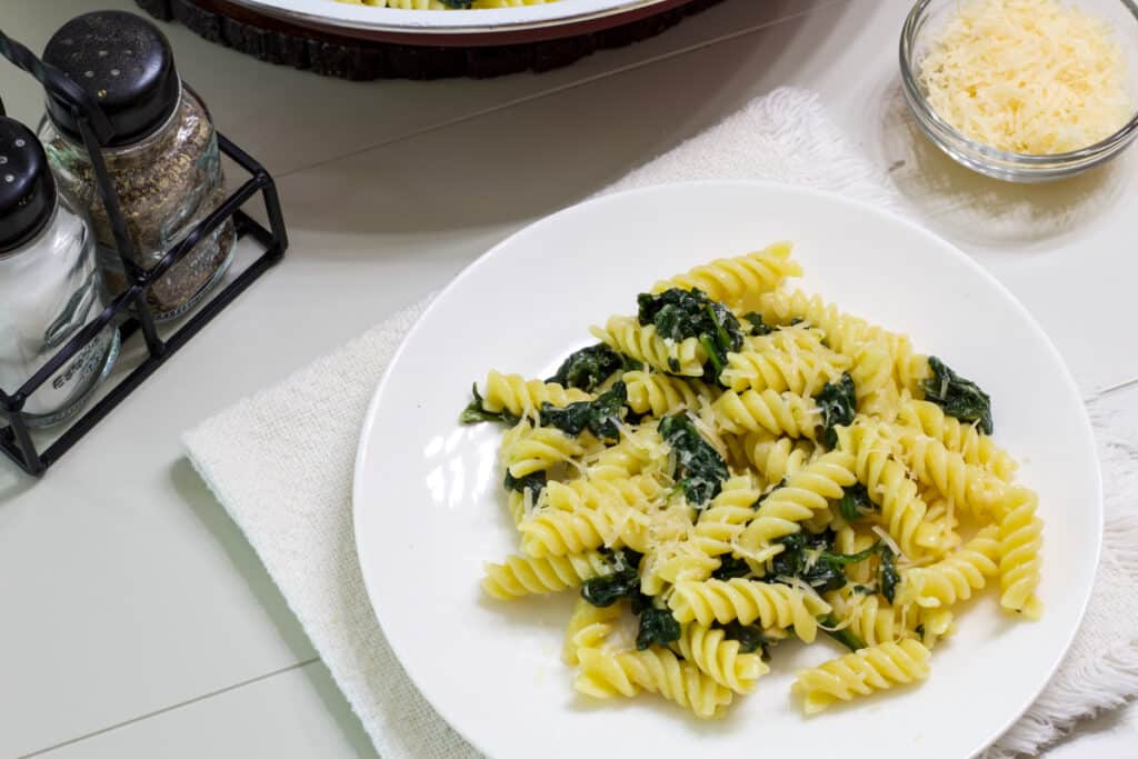 One serving of Easy Garlic Butter Pasta and Sautéed Spinach on a white plate, there is a small bowl of parmesan cheese and salt and pepper shakers near it.