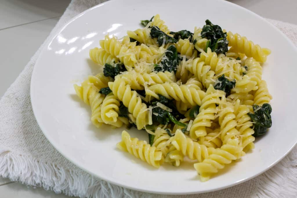 One serving of pasta with sauteed spinach on a white plate, the pasta has been sprinkled with parmesan cheese.