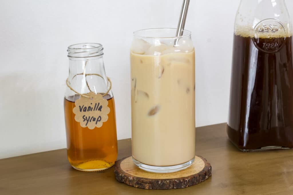 A glass of iced vanilla coffee and a bottle of vanilla syrup to the left of it and a bottle of black coffee to the right of it.