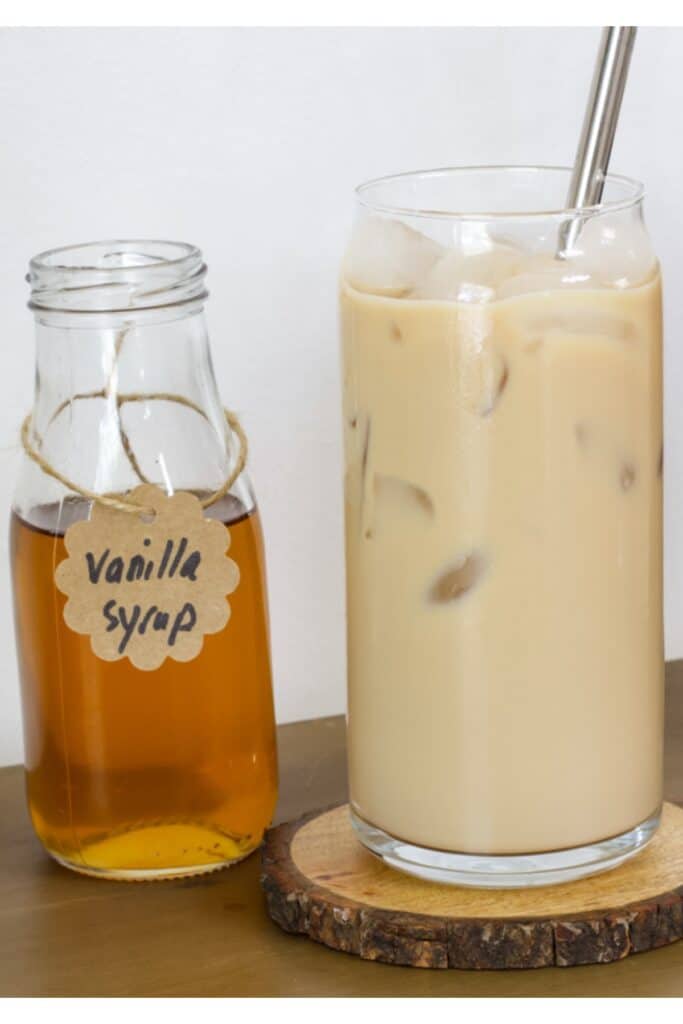 A small bottle of vanilla syrup sitting next to a tall glass of vanilla iced coffee.