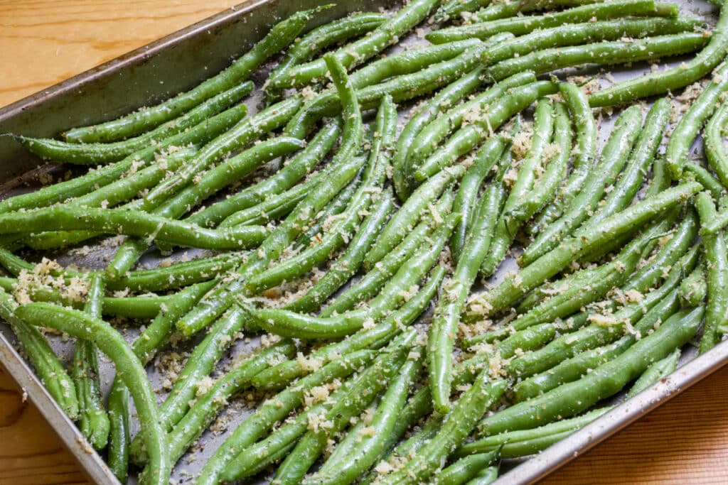 The green beans on the baking sheet after being coated with Parmesan cheese and bread crumbs and before being roasted.
