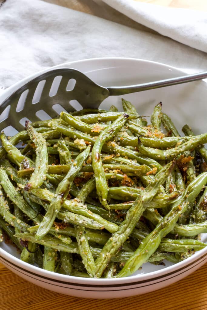 A bowl full of Roasted Green Beans with Paremasan Cheese, there is a slotted spoon in the bowl also.