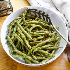 A large white serving bowl full of Roasted Green Beans with Paremasan Cheese and a slotted spoon is also in the bowl.