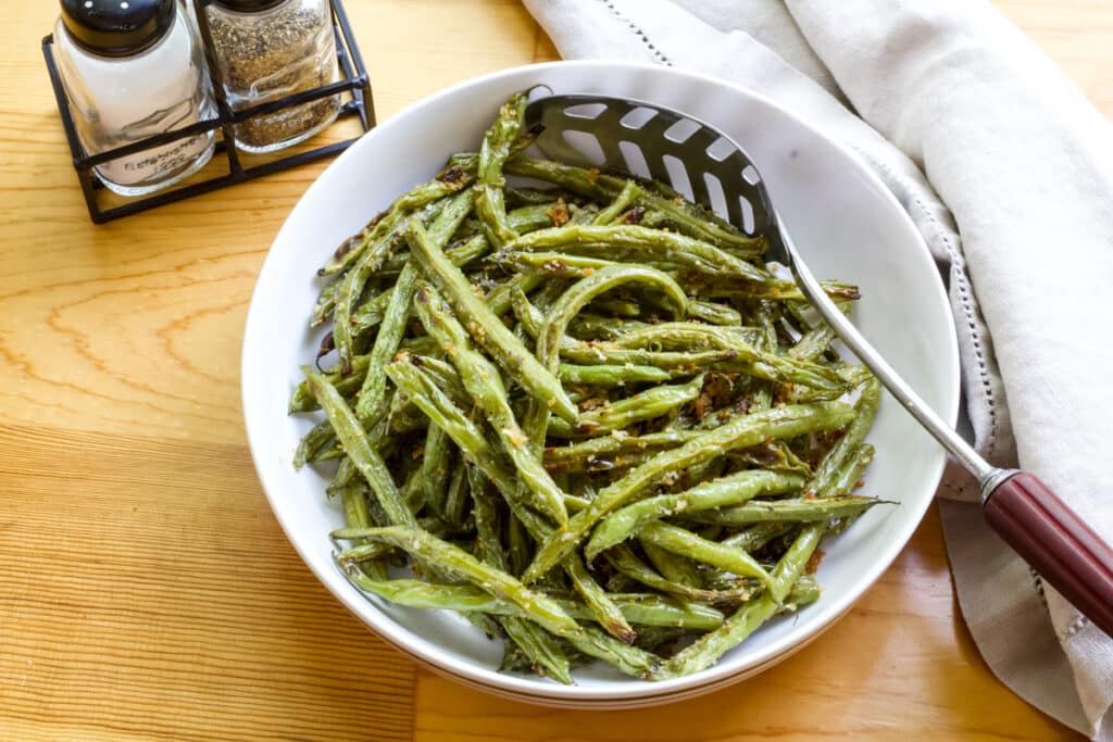 A serving bowl of Roasted Green Beans, there is a tan napkin to the right of it and salt and pepper shakers to the left of it.