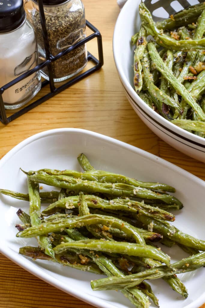 Close up of one serving of roasted green beans on a plate and the serving bowl and salt and pepper shakers are partially visible.