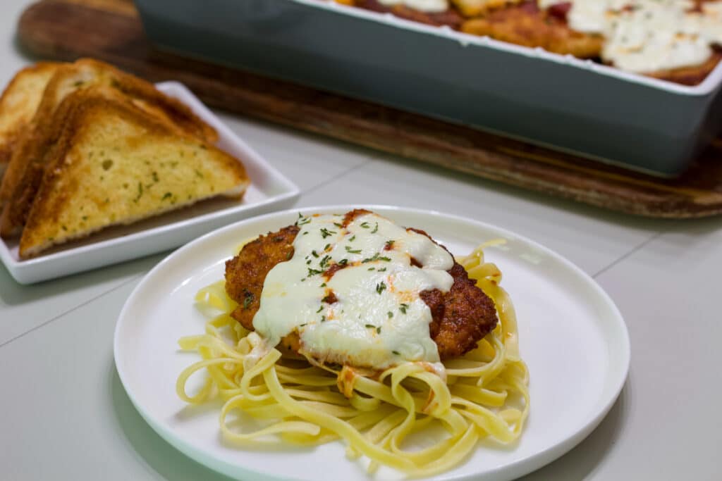 A plate with one serving of Crispy Chicken Parmesan, there is a plate of Texas toast in the background.