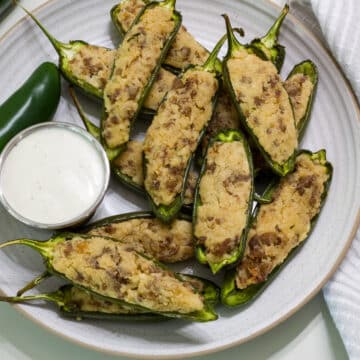 A round plate with several baked Stuffed Jalapenos on it, along side a bowl of ranch dressing.