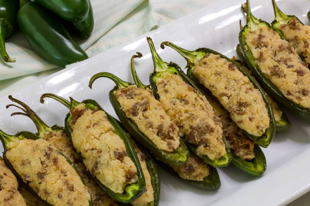 A plate with many cooked Stuffed Jalapenos, there are several fresh peppers in the background.