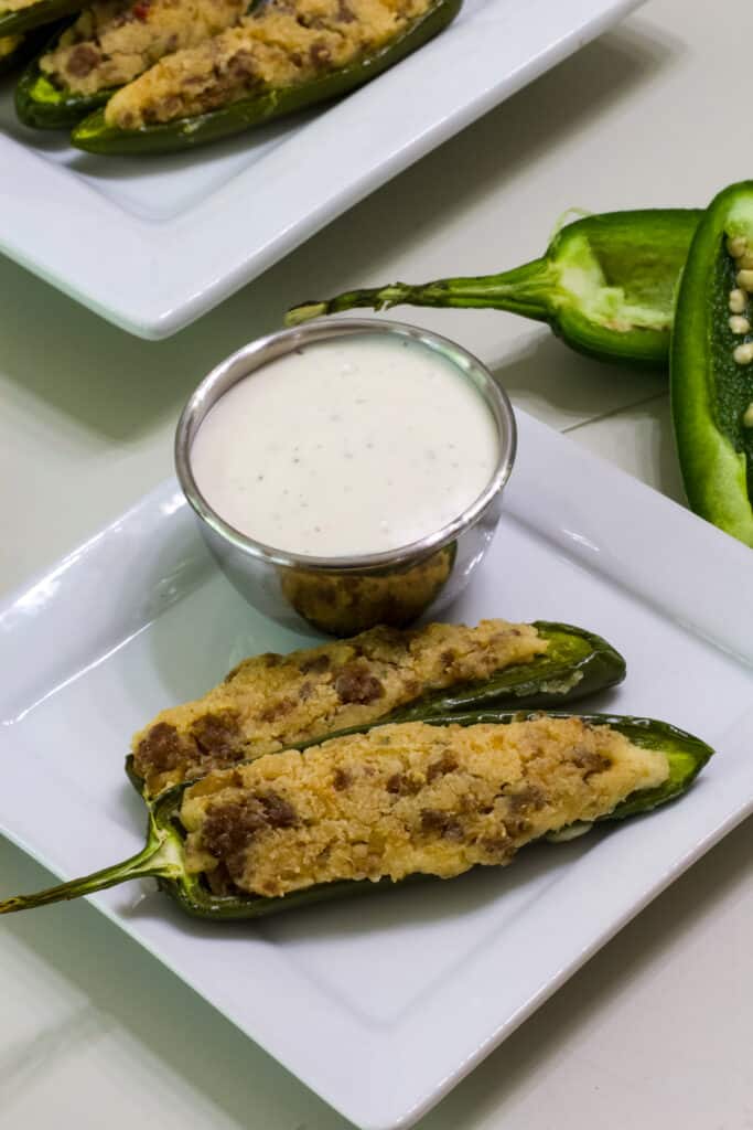 Two stuffed jalapeno peppers on a small white plate with a small bowl of ranch dressing.