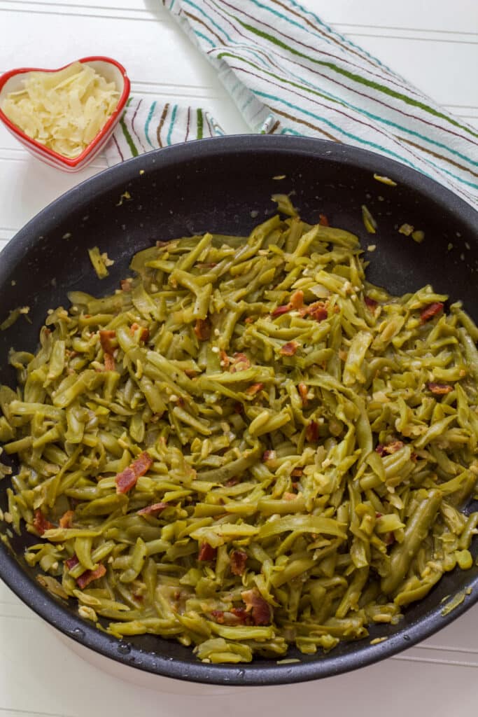 A large skillet wit the cooked green beans in it, there is a small heart shaped bowl above them that has the shaved Parmesan cheese in it.