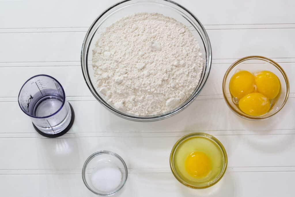A bowl of flour, egg yolks, whole egg, salt and a small measuring cup with water in it.