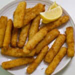 The feature image of several cooked fish stick on a plate with a coupele of lemon wedges.