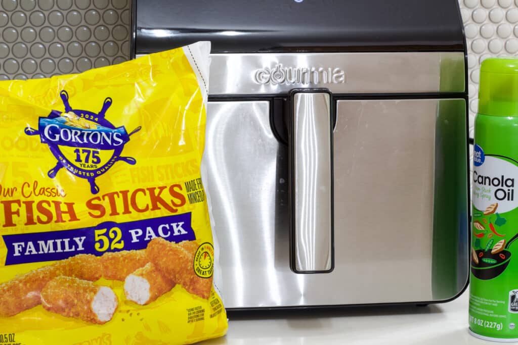 A family size bag of frozen fish sticks and can of non-stick cooking spray in front of the air fryer.