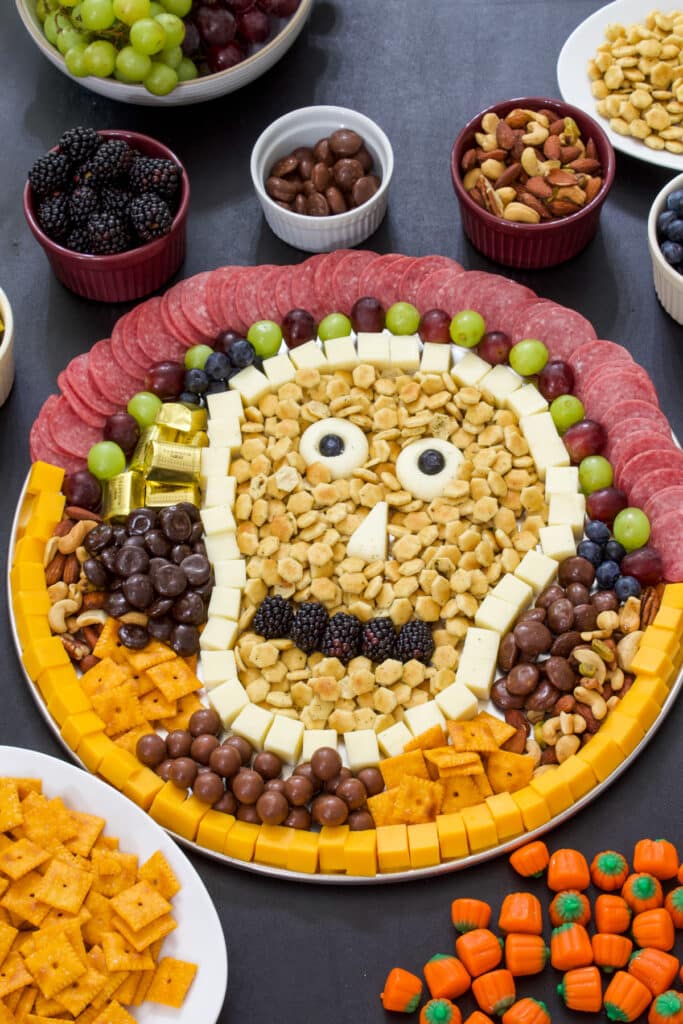 A large round pizza pan with meats, cheeses, fruits and candy in a skull shape, there are small bowls of additional ingredients around it.