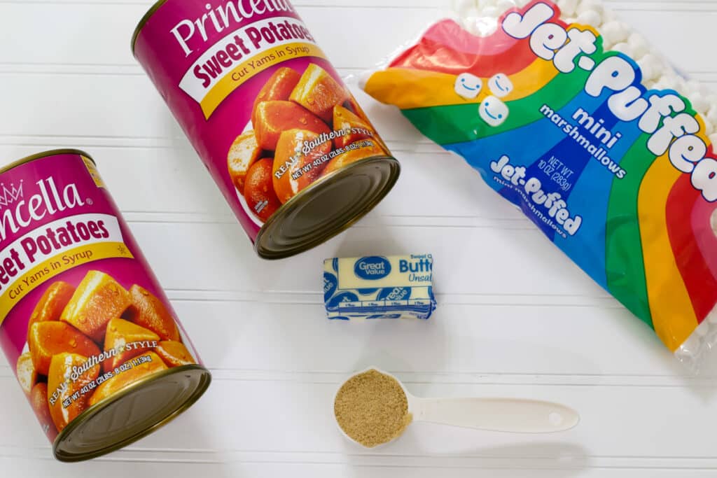Tow cans of sweet potatoes, a bag of miniature marshmallows, a half stick of butter and a measuring cup with light brown sugar.