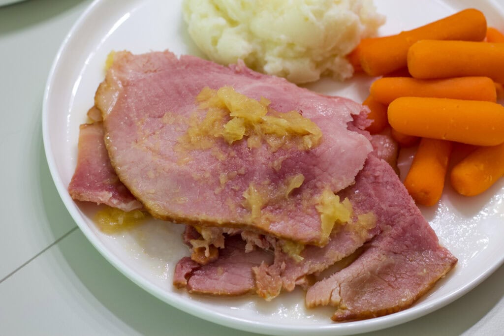 A pile of sliced ham, some carrots and mashed potatoes on a white plate.
