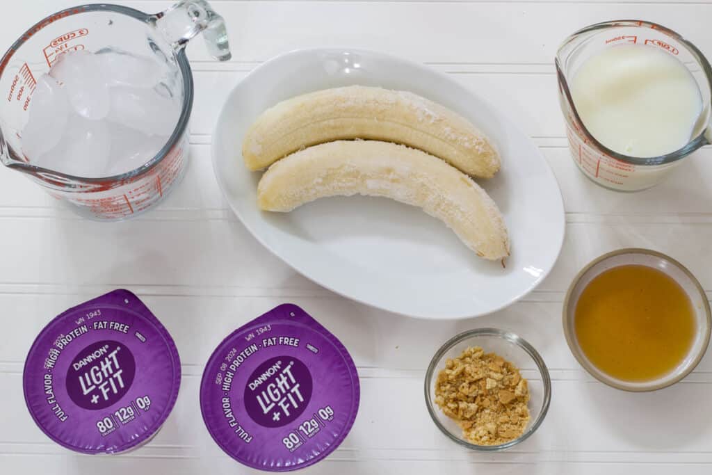 A cup of ice, two frozen bananas, cup of skim milk, two containers of yogurt, a small bowl of honey and a small bowl of graham cracker crumbs.