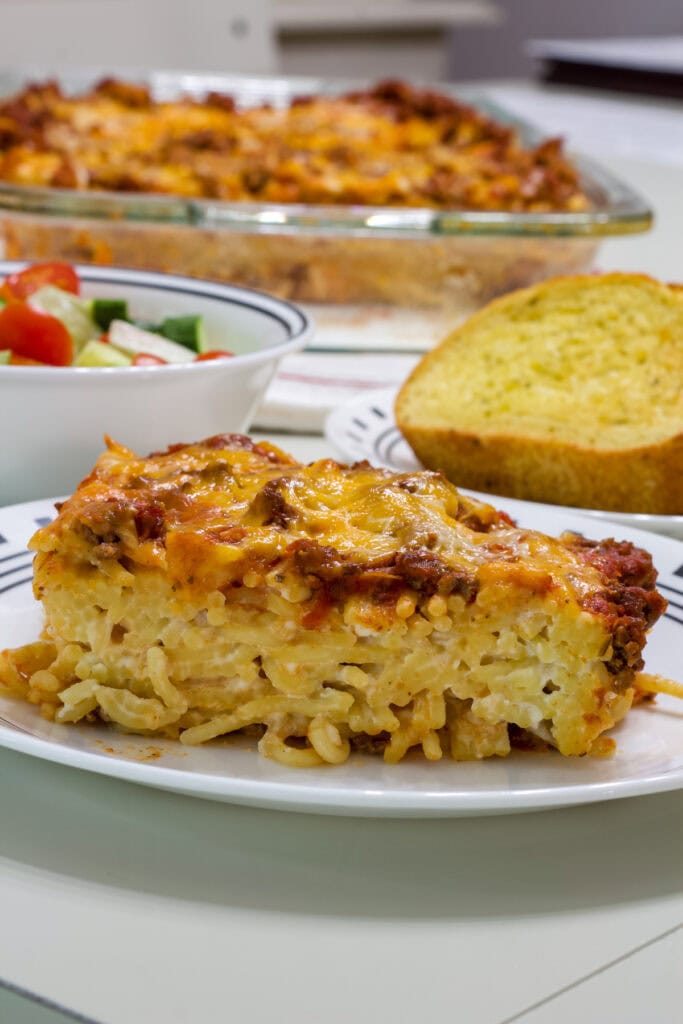 A plate with one serving of baked cream cheese spaghetti casserole in the foreground and a salad and garlic bread in the background.
