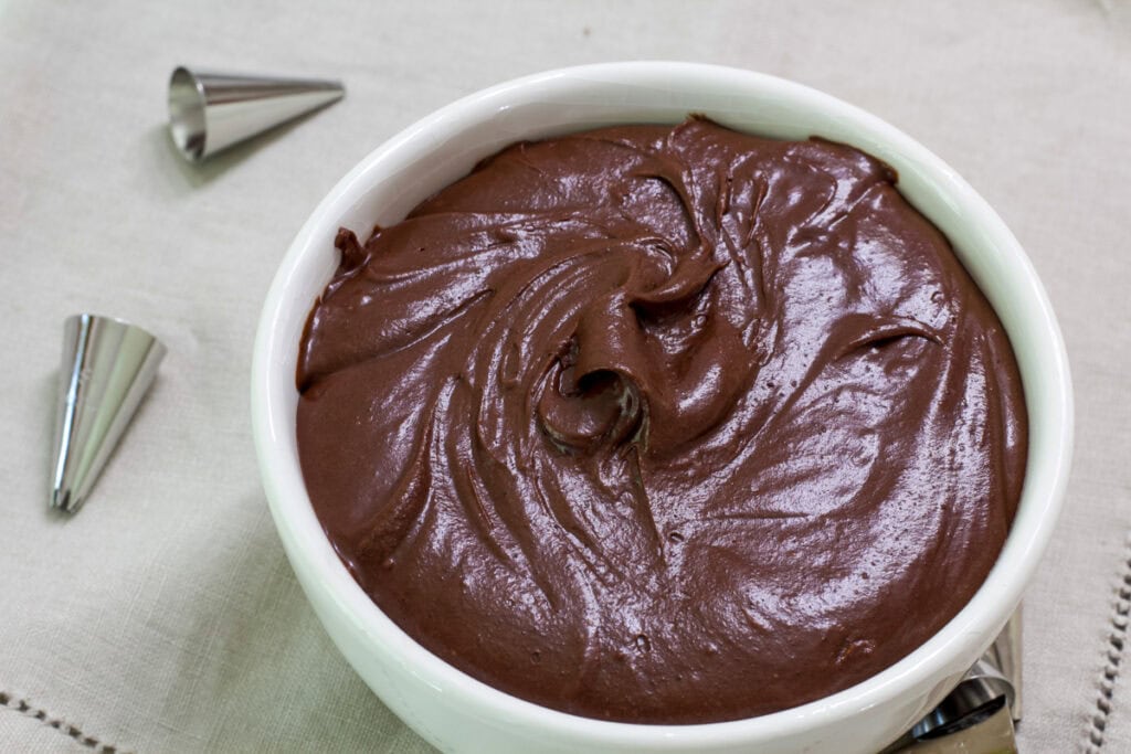 A white bowl of mocha frosting on a napkin with a couple of decorating tips next to it.