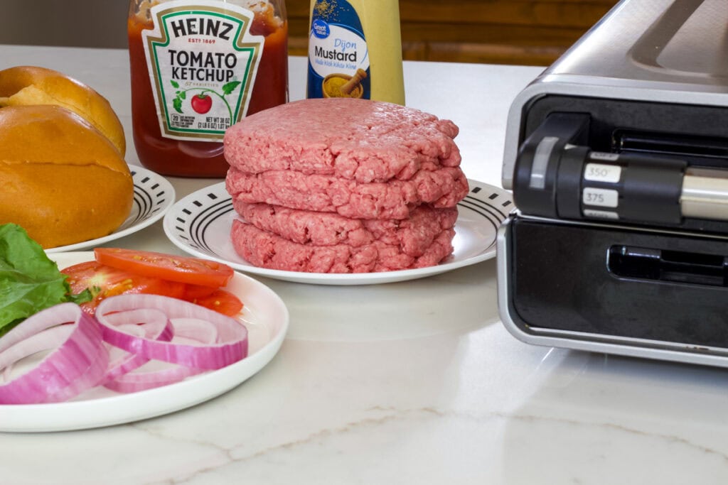 Four half pound burger on a plate, a plate with 2 hamburger buns, a plate with lettuce, tomatoes and red onion, and a George Foreman indoor grill sitting on the counter.