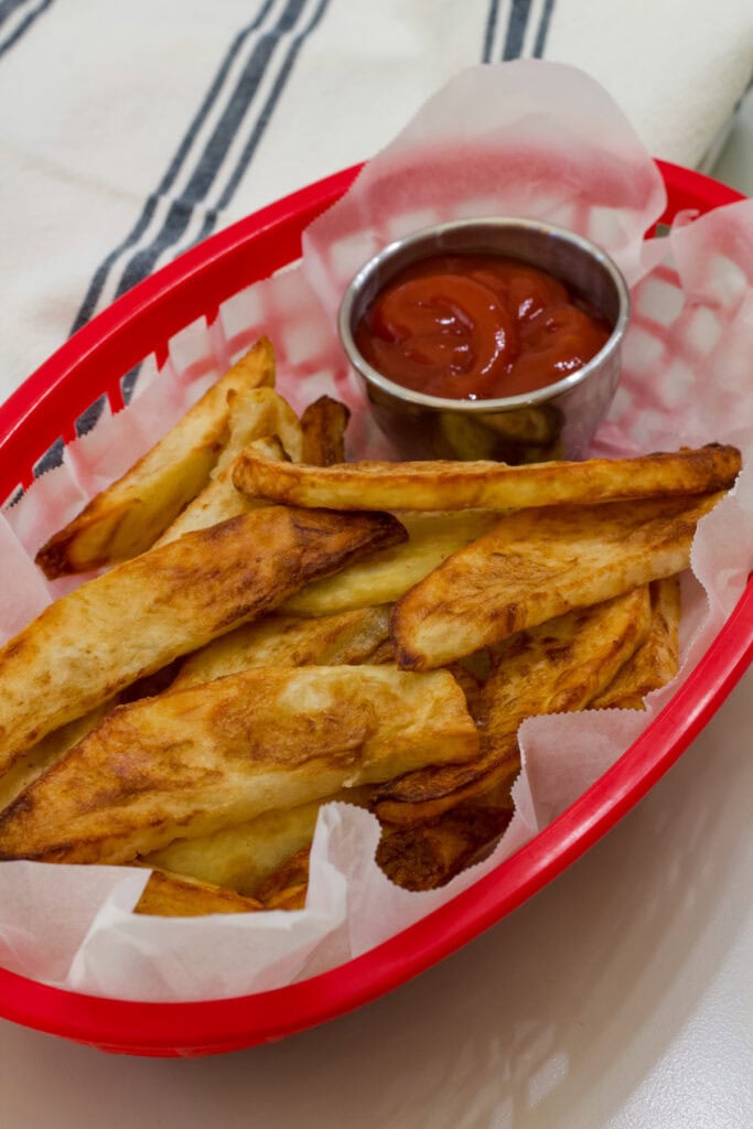 A red basket lined with parchment paper and filled with air fried steak fries and a small silver bowl of ketchup.