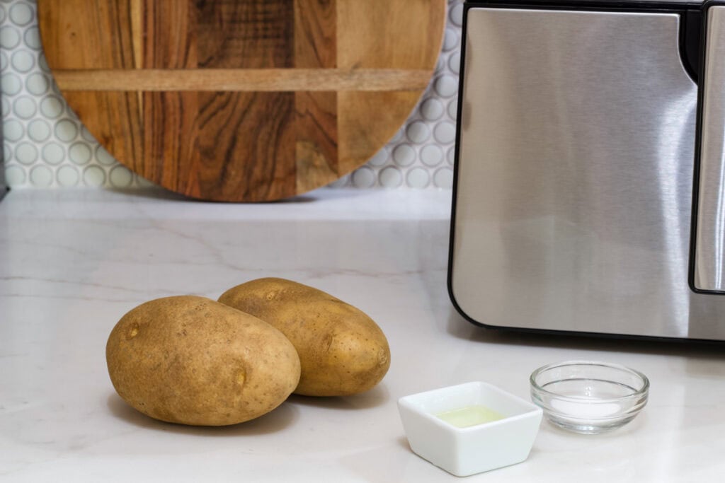 Two whole unpeeled potatoes, a small bowl of canola oil and a small bowl of salt sitting on the counter with the air fryer and a wooden cutting board.