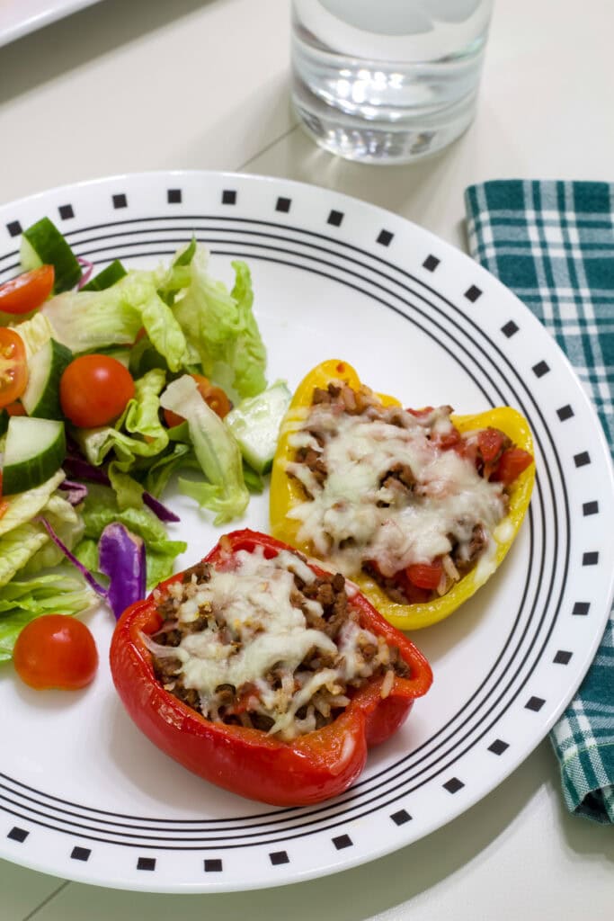 Two cooked stuffed peppers on a white plate with a salad, there is a glass of water in the background.