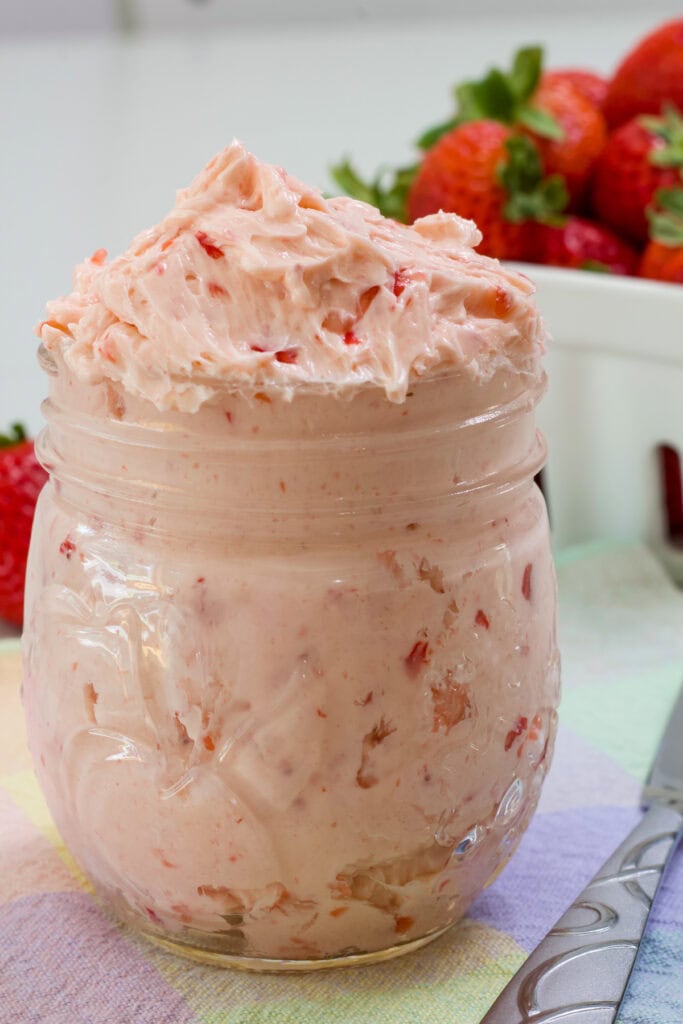 A small mason jar filled with Whipped Strawberry Honey Butter, there is a container of strawberries in the background.