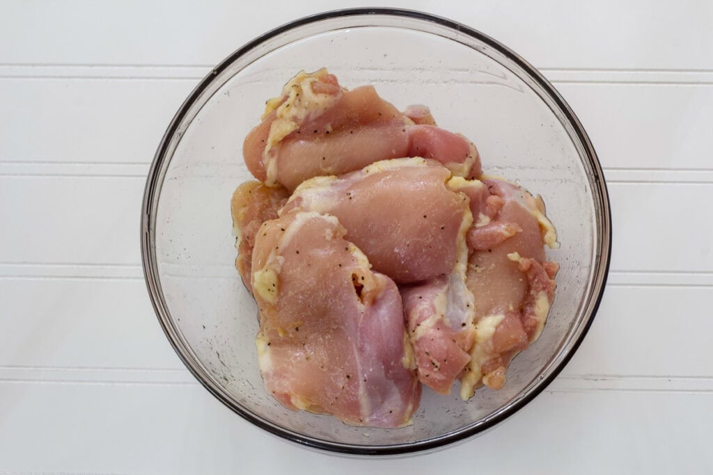 Five raw chicken thighs in a glass bowl after they have been tossed in oil and seasonings.
