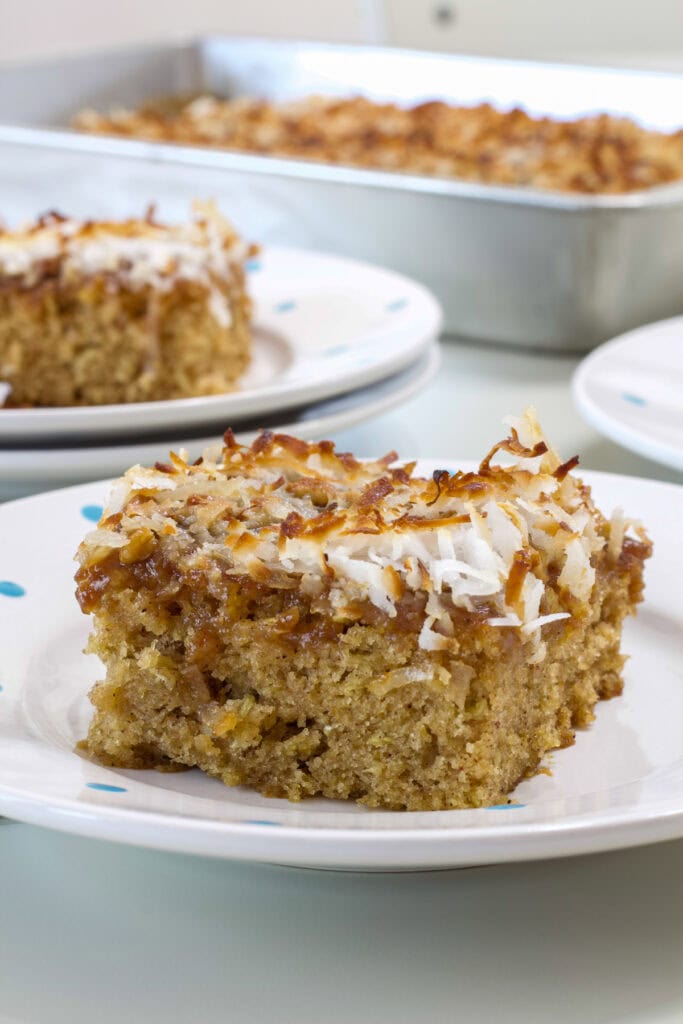 One piece of Oatmeal Cake with Broiled Coconut Topping on a plate, another piece and the rest of the cake is in the background.