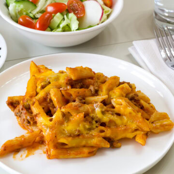 Side shot of one serving of Cheesy Ground Beef Pasta Bake on a white plate with a salad in the background.