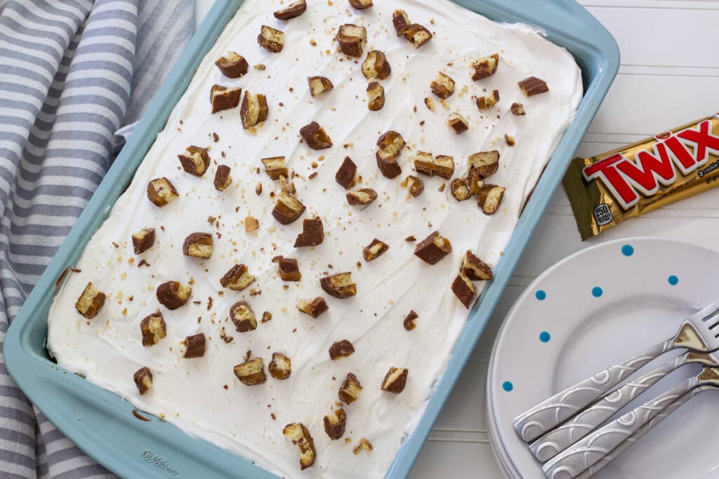 The entire uncut cake in a turquoise colored cake pan with some forks, plates and a Twix bar next to it.