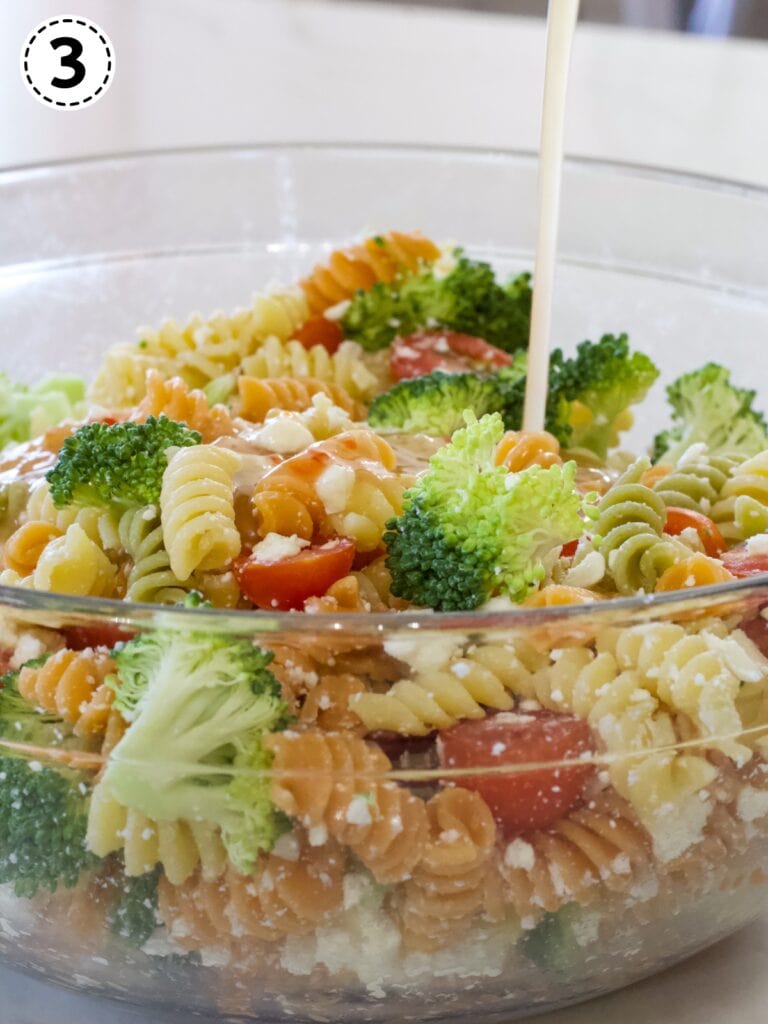 The pasta salad in a glass mixing bowl with the Italian dressing being poured onto it.