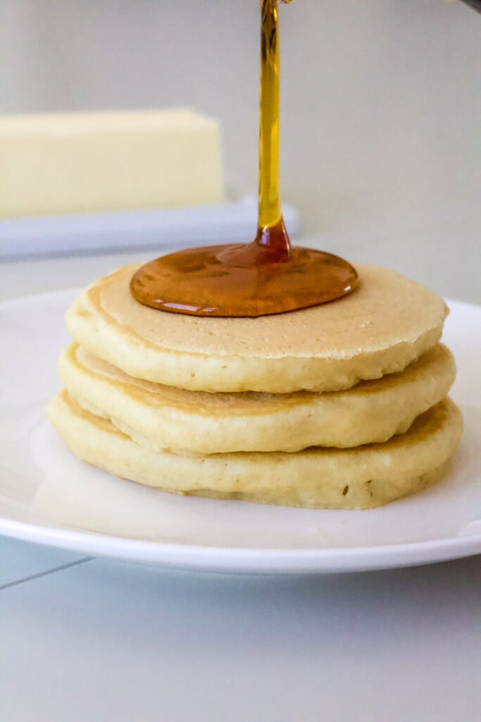 A short stack of three pancakes with syrup being poured down over them.