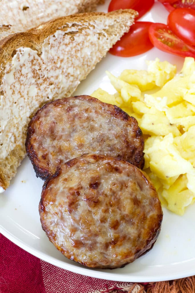 Two Air Fryer Sausage Patties on a plate with scrambled eggs, toast and sliced cherry tomatoes.