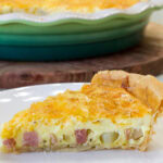 Feature image of the side view of one piece of Cheesy Ham & Potato Quiche on a plate.