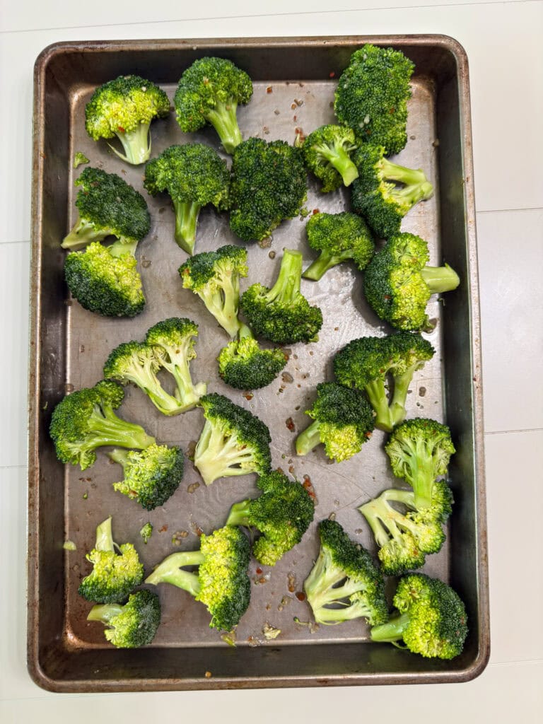 The raw broccoli on a sheet pan after it's been tossed in the dressing ready to go into the oven.