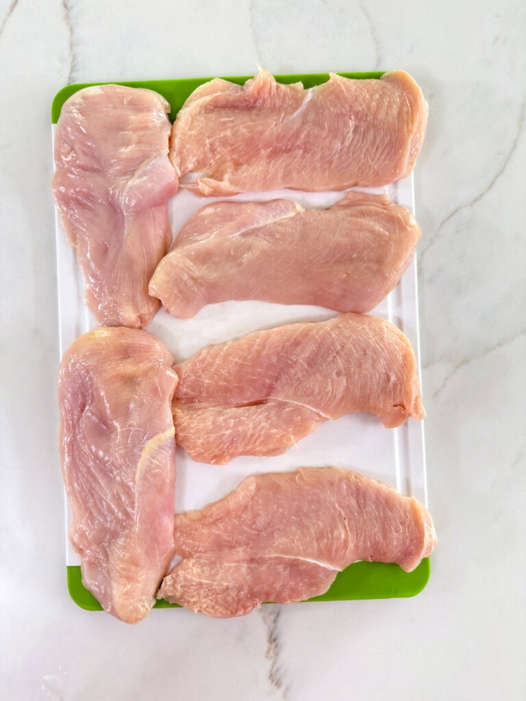 Six pieces of thinly sliced chicken breasts on a cutting board.