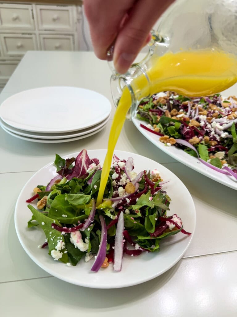 The orange vinaigrette being poured on to one serving of salad that's on a white plate.